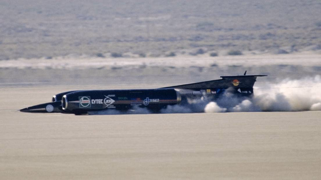 Jet-powered ThrustSSC speeding across a pale desert floor, trailing a cloud of dust